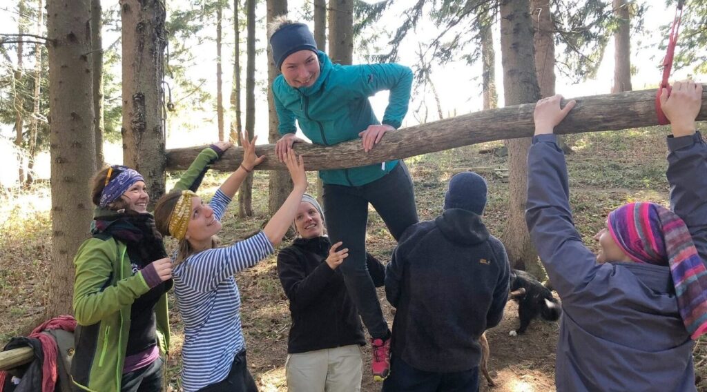 Gruppe sichert eine Teilnehmerin beim Übersteigen eines Baumstamms – Teamübung im Wald